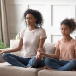 Happy mindful calm african american woman practicing yoga with healthy cute teenage daughter, teaching meditation in lotus pose and mudra gesture, relaxing together on couch in living room.