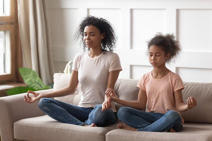Happy mindful calm african american woman practicing yoga with healthy cute teenage daughter, teaching meditation in lotus pose and mudra gesture, relaxing together on couch in living room.