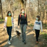 Mother with her children walking in public park with their mixed breed dog in time of corona virus outbreak. They wear protective mask and keeping social distance with other people as a prevention for spreading disease.